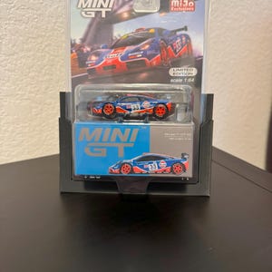 May include: A miniature McLaren F1 GTR #33 race car model in a clear plastic case. The car is blue, red, and orange with the number 33 on the side. The packaging includes the text "MINI GT" and "MiJo Exclusives". Scale 1:64.
