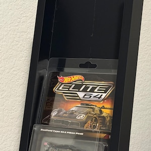 May include: Black display case holding a Hot Wheels Elite 64 Radford Type 62-2 Pikes Peak die-cast car. The car is in a clear plastic package with the Hot Wheels logo and car details visible.