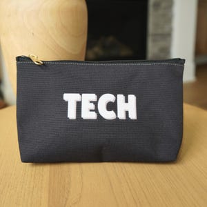 May include: A black canvas pouch with a gold zip and the word "TECH" in white block letters. The pouch is rectangular and appears to be made of a durable fabric. The background includes a wooden vase and a fireplace.