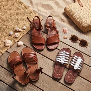 Op de afbeelding: Drie paar platte sandalen op een houten ondergrond. Eén paar bruine leren sandalen met enkelbandjes, één paar bruine leren sandalen met meerdere bandjes en één paar zilverkleurige metallic instapsandalen met meerdere bandjes en leren zolen.