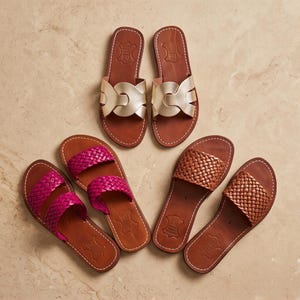 May include: Three pairs of flat sandals with brown leather soles. One pair features metallic gold wide straps, one pair has bright magenta woven straps, and one pair has tan woven straps. Each sandal has a stamped leather logo on the heel area.