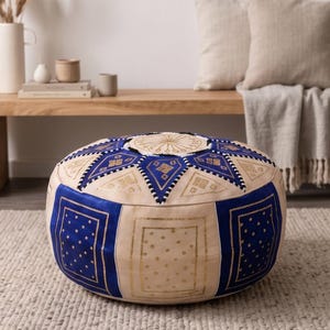 May include: Round, cream-colored pouf with blue and gold geometric patterns. The pouf has a circular top with gold embroidery and blue and cream panels. The design includes squares, triangles, and dotted patterns. It sits on a neutral-colored rug.