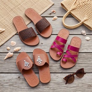 May include: Three pairs of sandals on a wooden surface: one pair with dark brown woven straps, one pair of bright pink sandals with crisscross straps and a toe loop, and one pair of tan leather slides featuring a silver triangular metal ornament.