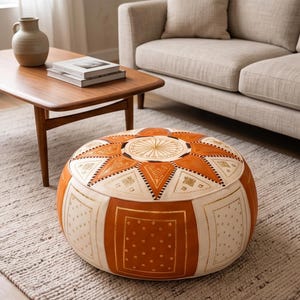 May include: A round, orange and cream-colored Moroccan pouf with geometric patterns. The pouf features a central starburst design and is surrounded by triangular and rectangular panels with gold accents. It sits on a neutral-toned rug, suggesting a living room setting.