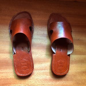 Puede incluir: Un par de sandalias de cuero marrón. Las sandalias tienen un diseño sencillo con una correa en la parte superior y una suela cosida. El cuero es de un color marrón rojizo cálido. Las sandalias se exhiben sobre una superficie de madera.