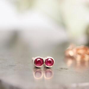 May include: A pair of stud earrings featuring round, bright red gemstones set in gold-coloured metal. The earrings are displayed on a reflective, grey surface, with a blurred background. The gemstones are the focal point of the image.