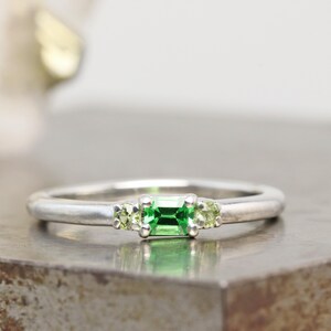 May include: A silver ring featuring a rectangular, emerald-cut green gemstone flanked by two smaller, round green gemstones. The ring has a simple, polished band and is set against a neutral background.