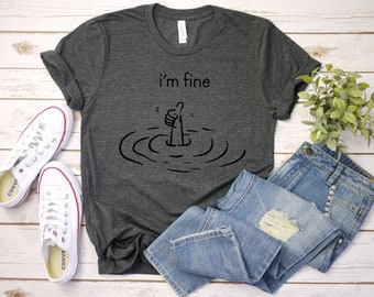 Könnte beinhalten: Ein dunkelgraues T-Shirt mit dem Text "i'm fine" und einer Cartoon-Hand, die einen Daumen hoch zeigt, aber die Hand ist in Wasser mit Wellen darum herum eingetaucht.