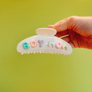May include: A cream-colored plastic hair claw clip featuring the text BOY MOM in raised, pastel-colored letters. The letters are individually colored in mint green, light pink, lavender, light blue, peach, and yellow, creating a soft contrast.