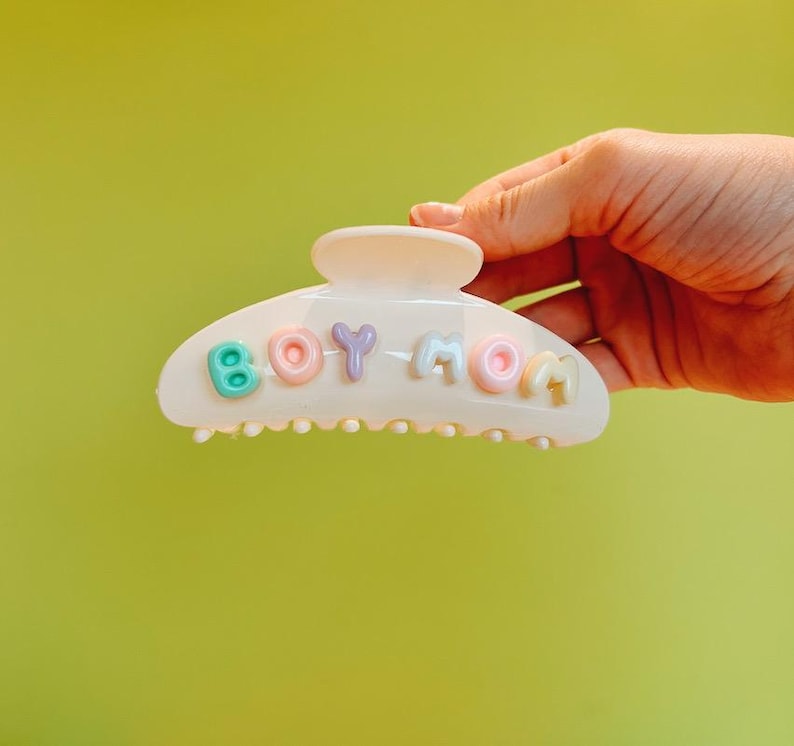 May include: A cream-colored plastic hair claw clip featuring the text BOY MOM in raised, pastel-colored letters. The letters are individually colored in mint green, light pink, lavender, light blue, peach, and yellow, creating a soft contrast.