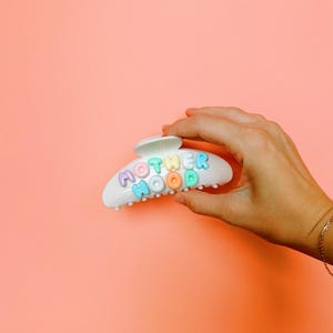 May include: A white plastic hair claw clip featuring the word MOTHERHOOD spelled out in raised, multi-colored pastel letters. The letters are arranged in two rows on the curved surface of the clip, showcasing shades of purple, blue, green, and orange.