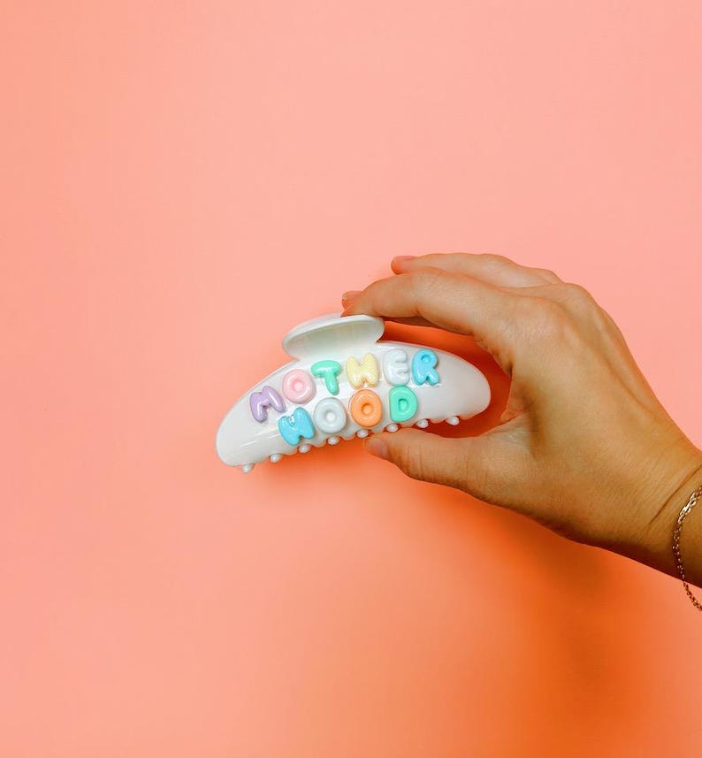 May include: A white plastic hair claw clip featuring the word MOTHERHOOD spelled out in raised, multi-colored pastel letters. The letters are arranged in two rows on the curved surface of the clip, showcasing shades of purple, blue, green, and orange.