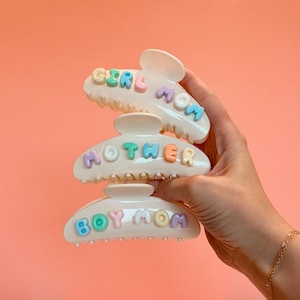 May include: Three off-white hair claw clips featuring pastel-colored, raised, bubble-style lettering. The top clip reads GIRL MOM, the middle clip reads MOTHER, and the bottom clip reads BOY MOM. Each letter is in a different shade of blue, pink, or yellow.
