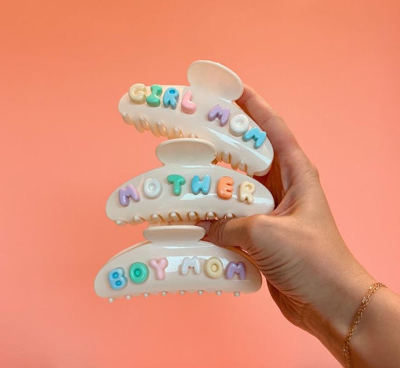 May include: Three off-white hair claw clips featuring pastel-colored, raised, bubble-style lettering. The top clip reads GIRL MOM, the middle clip reads MOTHER, and the bottom clip reads BOY MOM. Each letter is in a different shade of blue, pink, or yellow.