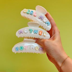 May include: Three white plastic hair claw clips stacked vertically. Each clip features raised, pastel-colored letters spelling out phrases. The top clip reads GIRL MOM, the middle clip reads MOTHER, and the bottom clip reads BOY MOM in various colors.