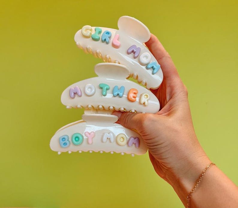 May include: Three white plastic hair claw clips stacked vertically. Each clip features raised, pastel-colored letters spelling out phrases. The top clip reads GIRL MOM, the middle clip reads MOTHER, and the bottom clip reads BOY MOM in various colors.