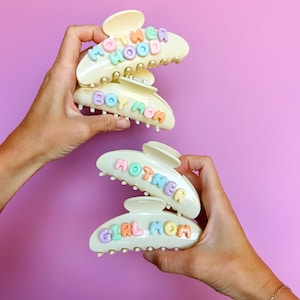 May include: Four cream-colored hair claw clips featuring pastel-colored, raised lettering. The top two clips display the words MOTHERHOOD and BOY MOM, while the bottom two clips display the words MOTHER and GIRL MOM in a rounded, bubbly font style.