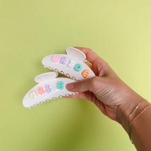May include: Two white hair claw clips with pastel-colored letters spelling "BRIDE" and "MRS B". The clips are held against a light green background. The letters are in shades of pink, orange, blue, and green.