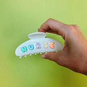 Puede incluir: Pinza para el pelo de pl&aacute;stico blanco con la palabra "NURSE" en letras de burbujas de colores. La pinza se sostiene sobre un fondo verde brillante.