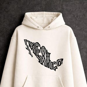 Puede incluir: Una sudadera con capucha de color crema que presenta un estampado gráfico negro del mapa de México. El mapa está relleno con patrones geométricos intrincados y la palabra MEXICO está integrada en el diseño. La sudadera tiene mangas largas.