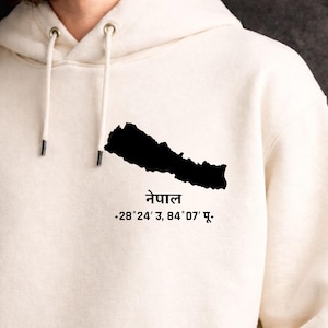 Puede incluir: Sudadera con capucha color crema con una silueta negra de Nepal y texto en idioma extranjero. La sudadera tiene una capucha con cordón y un bolsillo de canguro. El texto incluye la palabra "Nepal" y coordenadas.