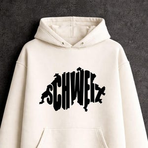 May include: A cream-colored hooded sweatshirt featuring a black graphic print on the chest. The graphic consists of the word SCHWEIZ stylized to form the shape of the map of Switzerland. The hoodie includes a front kangaroo pocket and long sleeves.