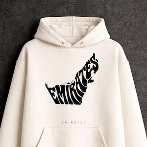 Puede incluir: Sudadera con capucha color crema con un gráfico negro del mapa de los Emiratos Árabes Unidos relleno con la palabra "EMIRATES". La palabra "EMIRATES" también está impresa debajo. Cuenta con capucha con cordones y bolsillo delantero.