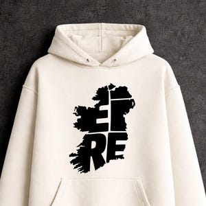 Może przedstawiać: Kremowa bluza z kapturem z czarnym nadrukiem graficznym przedstawiającym wyspę Irlandię. Wzór zawiera słowo EIRE zapisane dużymi, pogrubionymi, czarnymi wielkimi literami, które są wkomponowane w kształt mapy wyspy.