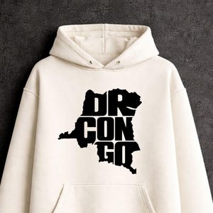 Puede incluir: Una sudadera con capucha de color crema que presenta un estampado gráfico negro en el pecho. El gráfico es una silueta del mapa de la República Democrática del Congo con el texto DR CON GO en letras mayúsculas negras y negritas.