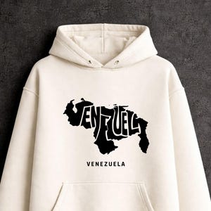 Puede incluir: Una sudadera con capucha de color crema que presenta un estampado gráfico negro del mapa de Venezuela. El mapa contiene el texto estilizado "VEN" y "UEL" integrado en su forma, con la palabra "VENEZUELA" impresa en letras mayúsculas negras debajo.