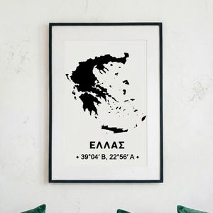 May include: A framed print showing a black silhouette of Greece on a white background. The word "ΕΛΛΑΣ" is centered below the map, with coordinates 39°04'B, 22°56'A below. The frame is black.