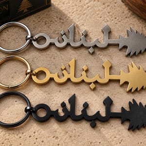 May include: Three metal keychains in silver, gold, and black tones. Each keychain features a circular ring attached to a flat, stylized Arabic script design that transitions into a pointed, triangular cedar tree shape at the end.