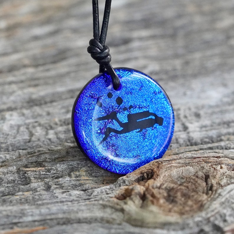 May include: A blue glass pendant necklace featuring a black silhouette of a scuba diver. The pendant has a textured surface and is on a black leather cord.