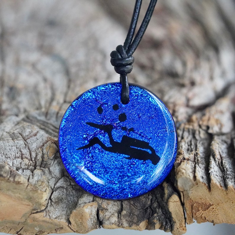 May include: A blue glass pendant necklace featuring a black silhouette of a person jumping rope. The pendant is round and has a textured surface. The necklace is made with a black cord.