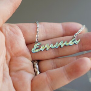 May include: A silver chain necklace with a cursive nameplate pendant that says "Emma" in a light blue and yellow color scheme.