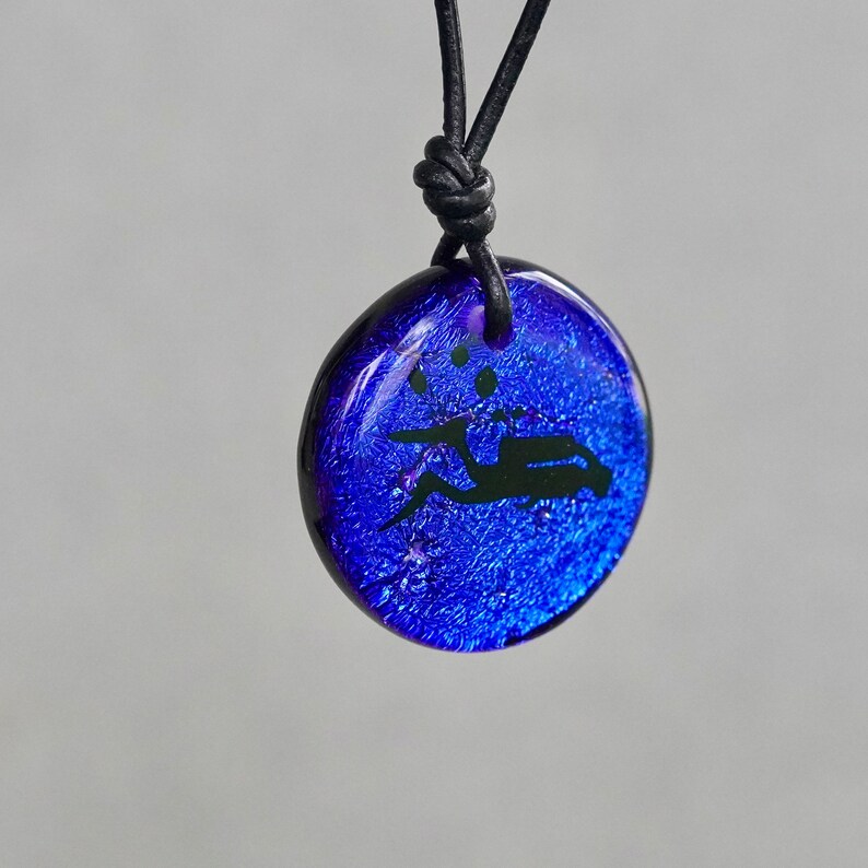 May include: A blue glass pendant necklace featuring a black silhouette of a person riding a scooter. The pendant has a textured, sparkly surface.