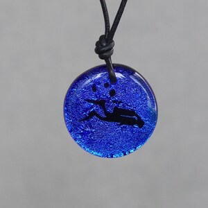 May include: A blue glass pendant necklace with a black leather cord. The pendant features a black silhouette of a diver with bubbles.
