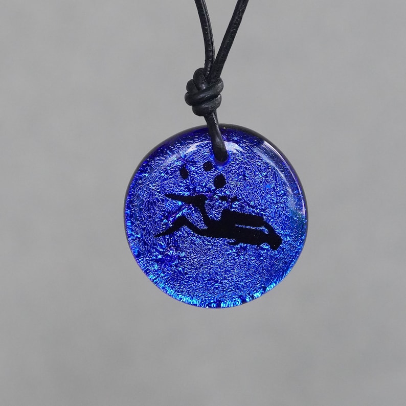 May include: A blue glass pendant necklace with a black leather cord. The pendant features a black silhouette of a diver with bubbles.
