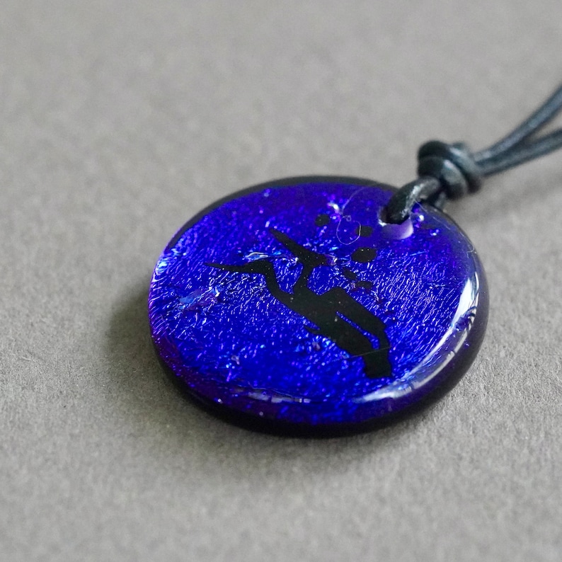 May include: A blue glass pendant with a black hummingbird silhouette, hanging from a black leather cord.