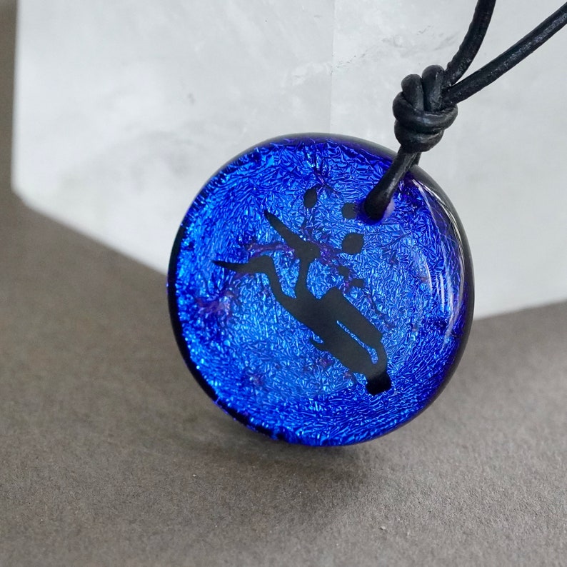 May include: A blue glass pendant featuring a black silhouette of a tree branch with leaves. The pendant is on a black leather cord.