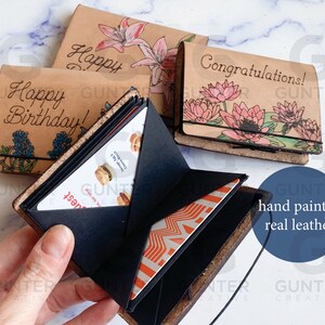 May include: Three leather wallets with hand-painted designs. The wallets are open to show the interior, which has multiple card slots. The wallets are decorated with flowers and the text "Happy Birthday!" and "Congratulations!"