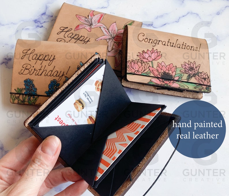 May include: Three leather wallets with hand-painted designs. The wallets are open to show the interior, which has multiple card slots. The wallets are decorated with flowers and the text "Happy Birthday!" and "Congratulations!"
