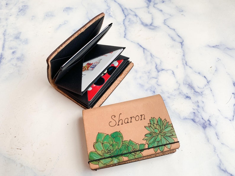 May include: A brown leather wallet with a black interior and a floral design. The wallet is open and has a card slot and a cash pocket. The wallet is personalized with the name "Sharon".