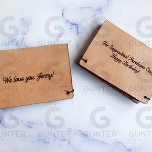May include: Two brown leather wallets with engraved text. The first wallet says "We love you Jenny!" The second wallet says "For Impractical Purchases Only Happy Birthday!"