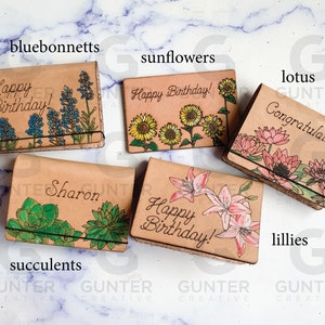 May include: Five leather card holders with different floral designs. The designs include bluebonnets, sunflowers, lotuses, lilies, and succulents. Each card holder has a different inscription, including "Happy Birthday!" and "Congratulations!"