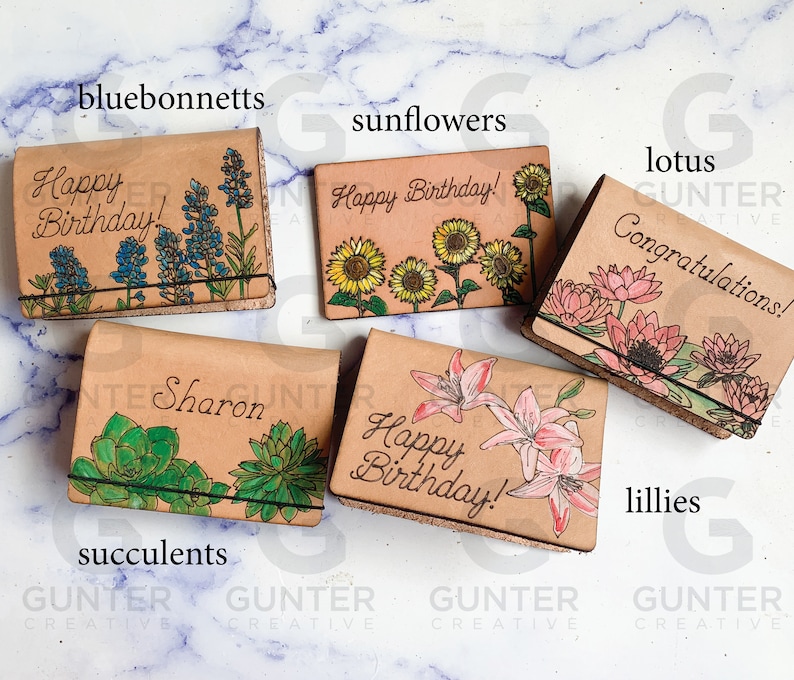 May include: Five leather card holders with different floral designs. The designs include bluebonnets, sunflowers, lotuses, lilies, and succulents. Each card holder has a different inscription, including "Happy Birthday!" and "Congratulations!"
