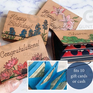 May include: Four leather gift card holders with different floral designs. The holders are open to show the blue card slots inside. The text on the holders reads "Happy Birthday!", "Happy Day!", "Congratulations!" and "Sharon". The text "fits 10 gift cards or cash" is also visible.