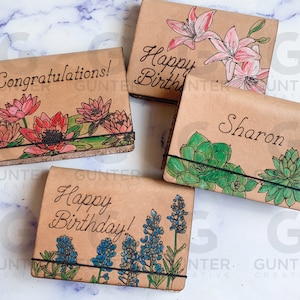 May include: Four small leather journals with different floral designs and personalized messages. The journals have black elastic bands and are made of brown leather. The messages include "Congratulations!", "Happy Birthday!", "Happy Birthday!" and "Sharon".