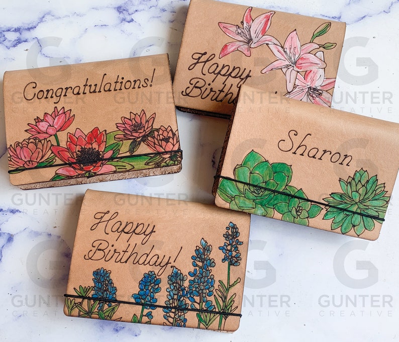 May include: Four small leather journals with different floral designs and personalized messages. The journals have black elastic bands and are made of brown leather. The messages include "Congratulations!", "Happy Birthday!", "Happy Birthday!" and "Sharon".