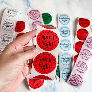 May include: Multiple rolls of circular adhesive stickers in red, pink, blue, and green. The stickers feature black text including Smith Elementary, Spirit Night!, and Fundraising Night, with specific event details like Sonic @ Louetta and times.